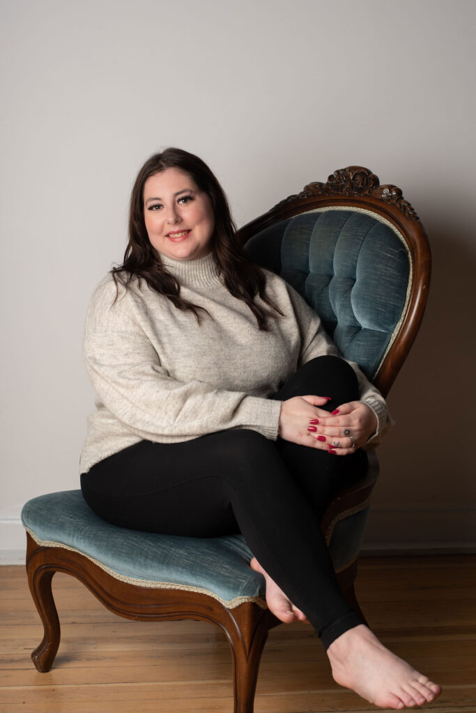 woman in sweater sitting in a ornate chair slighting hugging up one knee smiling at camera