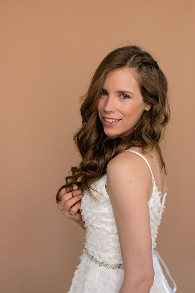 Bride looking over shoulder, gently holding some of her hair, with a soft smile.