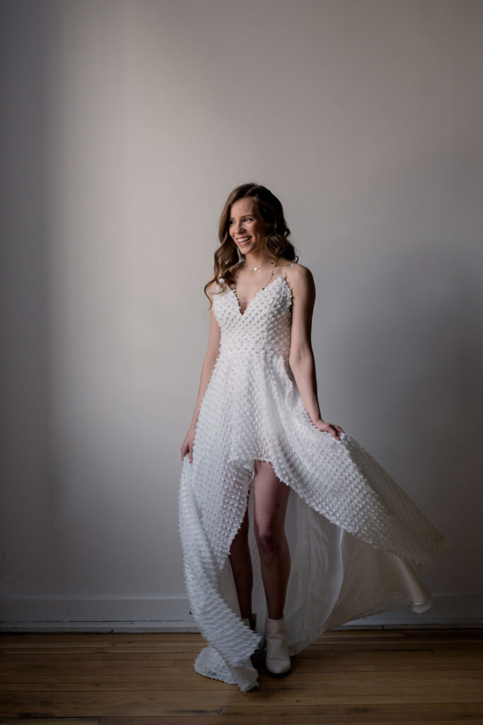 Bride wearing a high-low hem dress, in the middle of twirling her skirt, looking out the window and laughing.