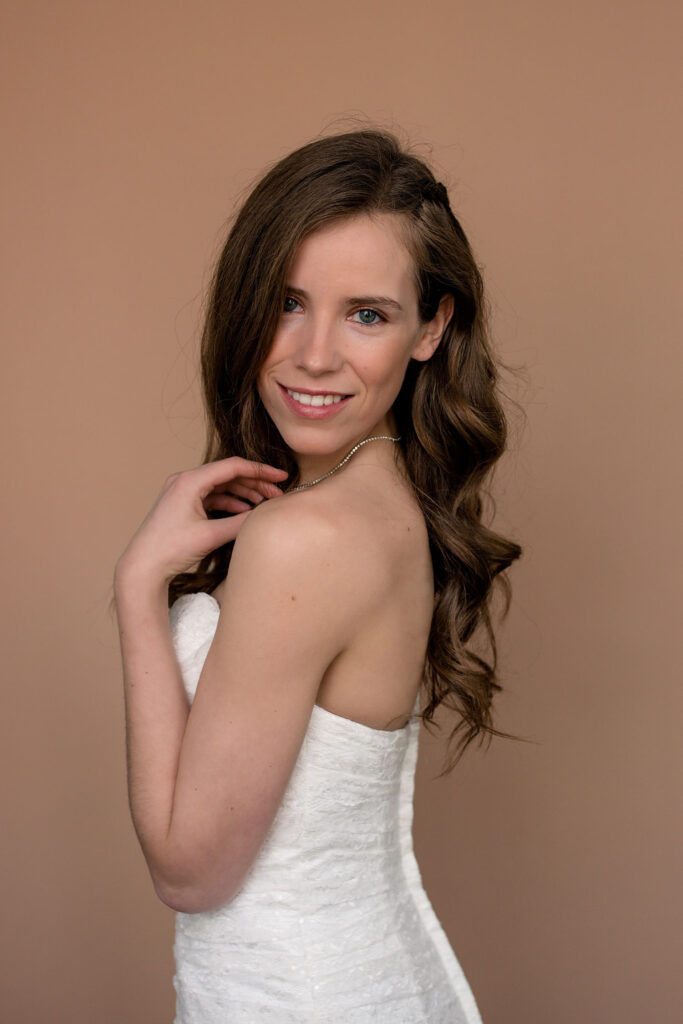 Bride looking over shoulder at camera with a soft smile, gently touching her collarbone.