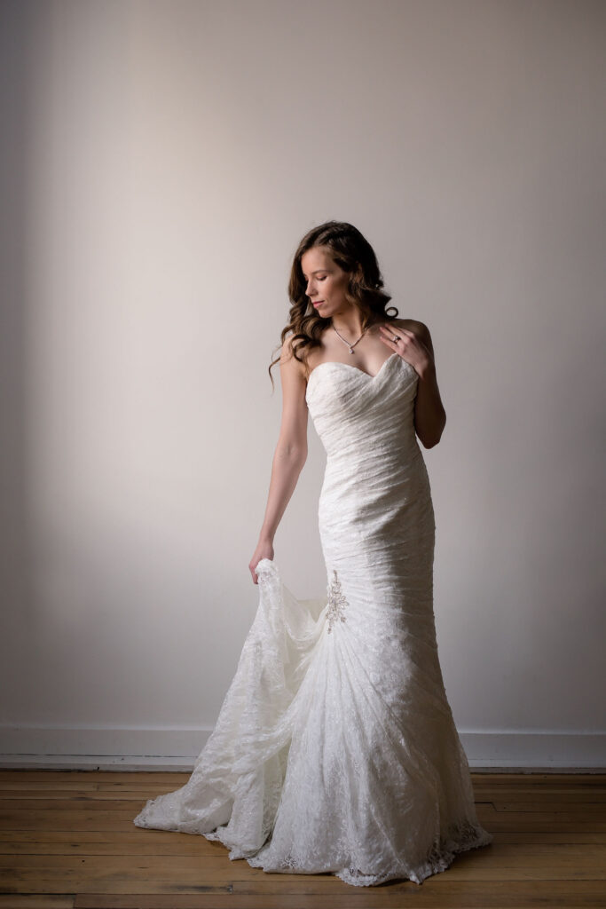 Bride looking down her shoulder while holding the train of her dress, opposite hand gently touching her collarbone.