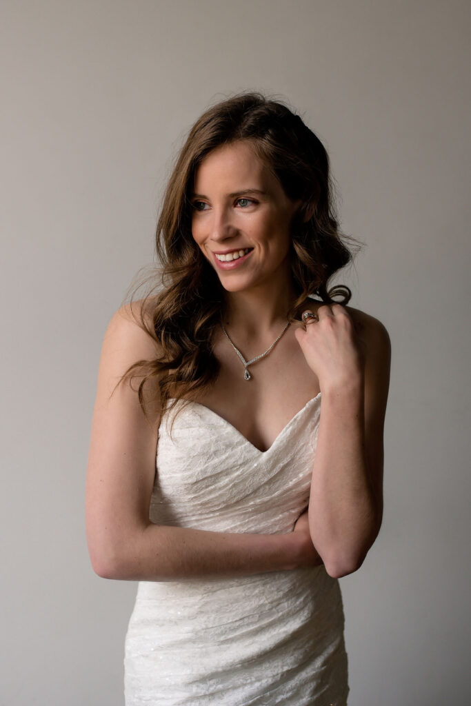 Bride smiling and looking off camera, one arm wrapped around her waist, the other gently brushing her collarbone