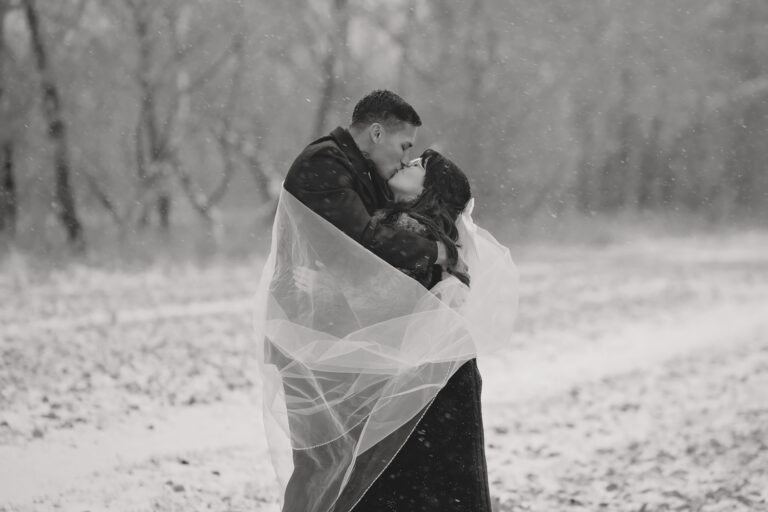 bride and groom kissing in the snow with her veil wrapped around them